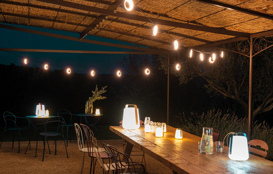 Fermob HOOPIK outdoor string lights L1200 cm hanging above an outdoor restaurant setting, creating warm ambient lighting.
