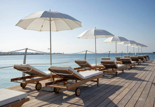 Fermob IBIZA teak outdoor sunlounger with a slatted design, placed on a beach with a wooden frame for relaxing in the sun.