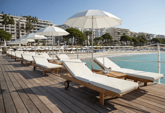 Fermob IBIZA teak outdoor sunlounger with a slatted design, placed on a beach with a clear sky and sandy surroundings.