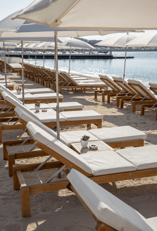 Fermob IBIZA teak sunlounger with wooden frame on a beach setting, outdoor sunbed for relaxing in the sun.